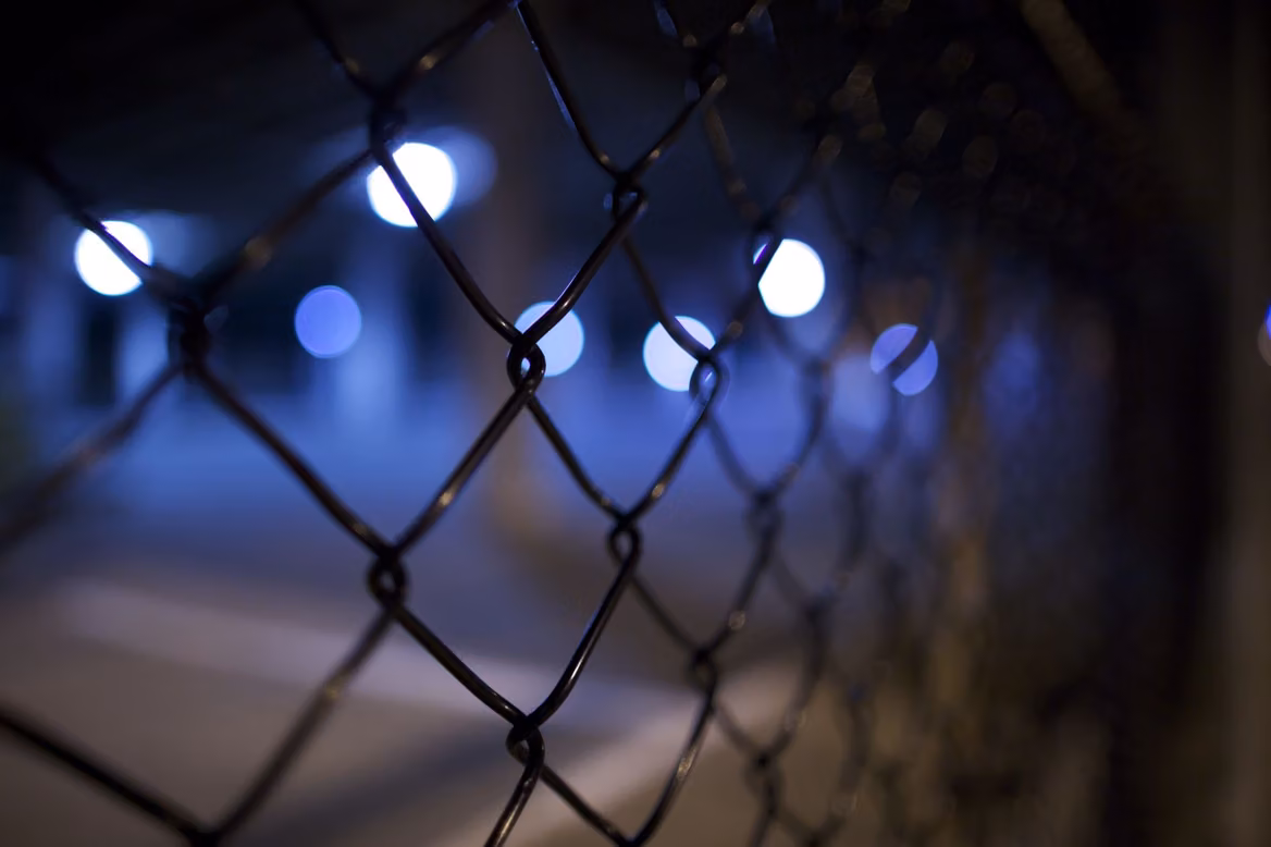 Close view of a chain-link fence at dusk, with blurred blue lights behind it.
