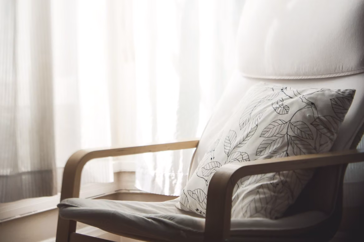 An empty armchair with a cushion beside a window hung with sheer white curtains in a softly lit room.