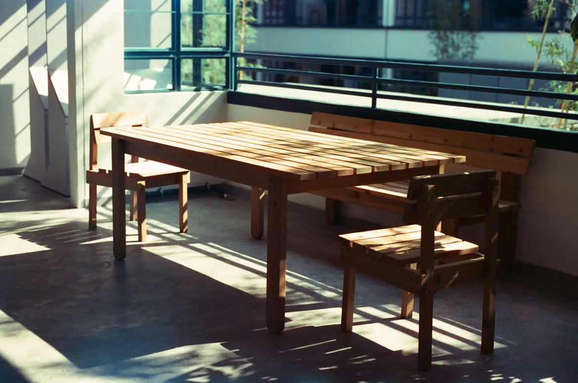 A wooden table, a bench and two chairs in a quiet, sunlit room, with long shadows across a concrete floor and windows looking out onto trees.